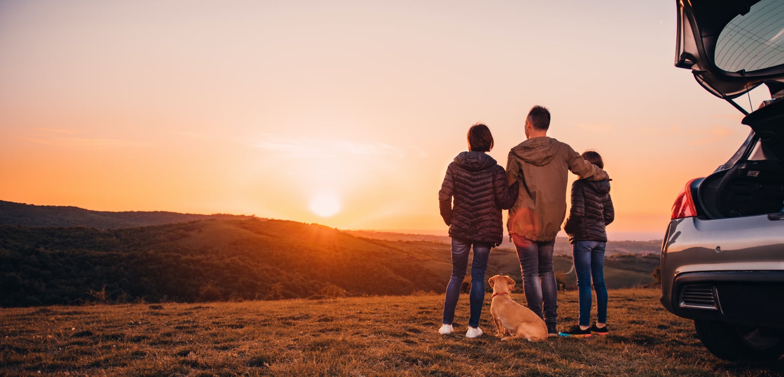 family standing outside of rental car watching sunset over hills