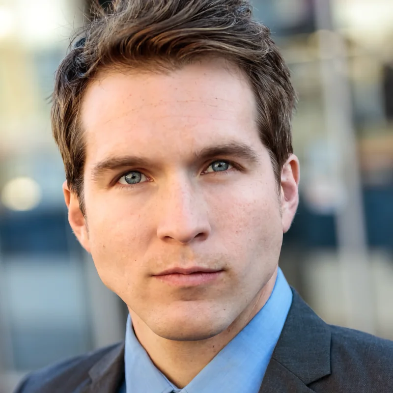 A close-up portrait of Dade Elza, a staff member, wearing a dark suit jacket and a blue shirt, looking directly at the camera with a focused expression.